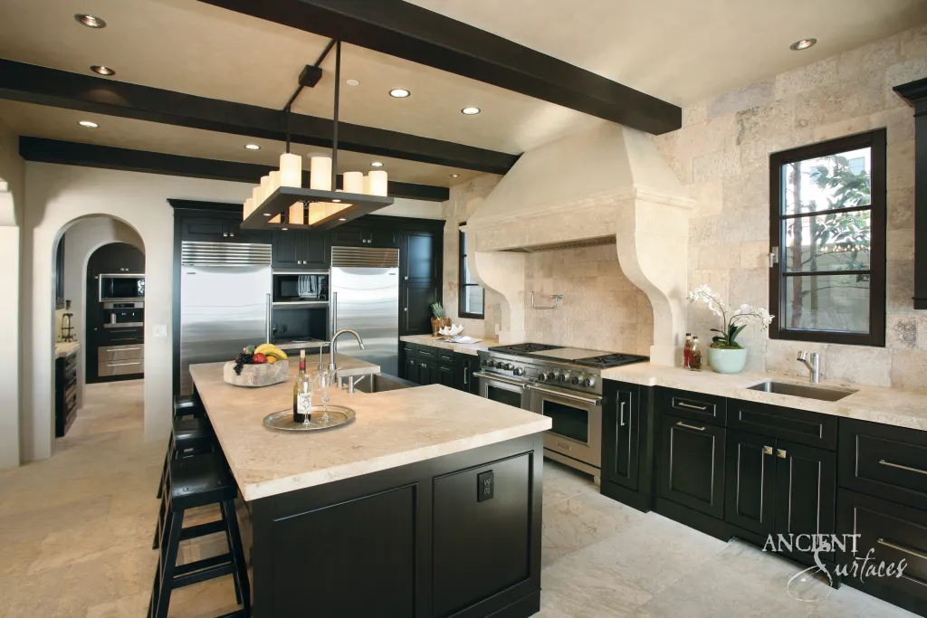 Limestone kitchen hood with scrolled brackets and limestone countertops over dark cabinetry beneath timber beams