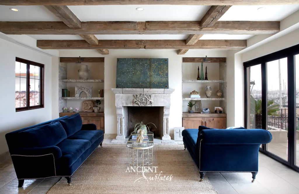 Carved antique French limestone mantel with a central cartouche, flanked by shelved alcoves in a beamed living room