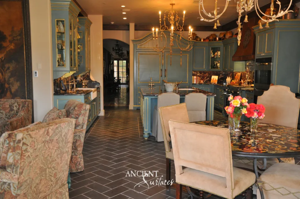 Herringbone reclaimed French limestone floor running from the dining area into a teal kitchen, beside a marble inlaid dining table set with roses