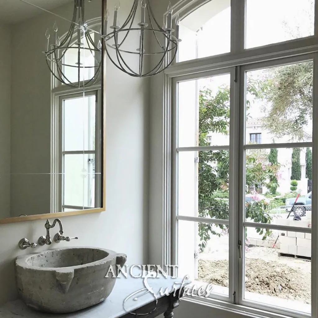 Carved limestone basin with a pouring lip on a vanity beneath a wall mounted faucet beside a tall bathroom window